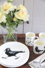 jackdaw plate in foreground with jackdaw mug and vase of flowers behind