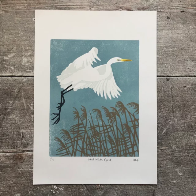 white bird with black legs flying over brown grasses against dull blue background