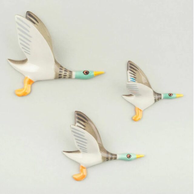 Three ceramic ducks with yellow legs and feet flying across a wall