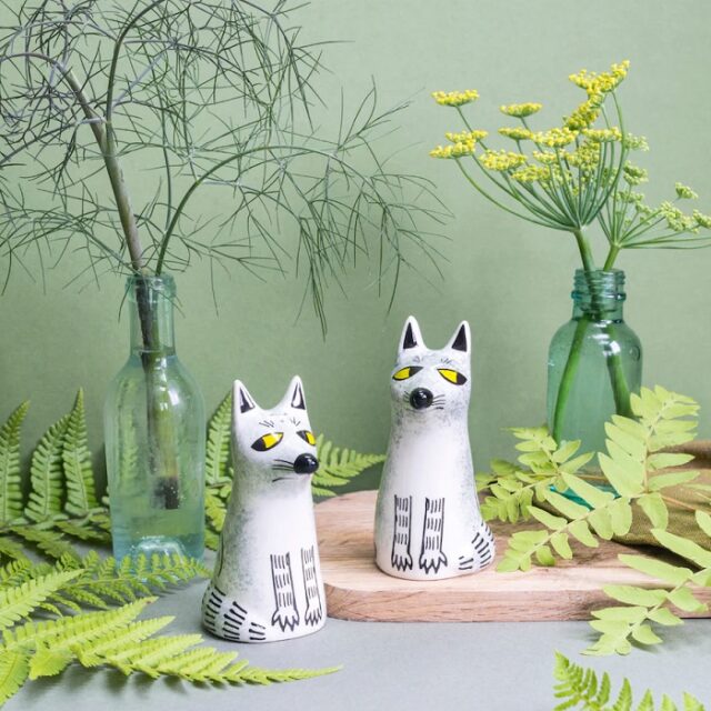 Two grey wolf salt and pepper shakers are shown between green bottles with ferns and grasses around
