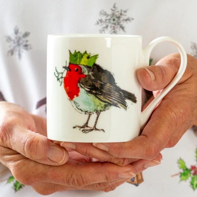A woman's hands holding a white mug with a robin on and the robin is wearing a green paper hat