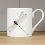 Dragonfly mug by holly lasseter shows a water colour drawing of a dragonfly in grey and black on a white mug