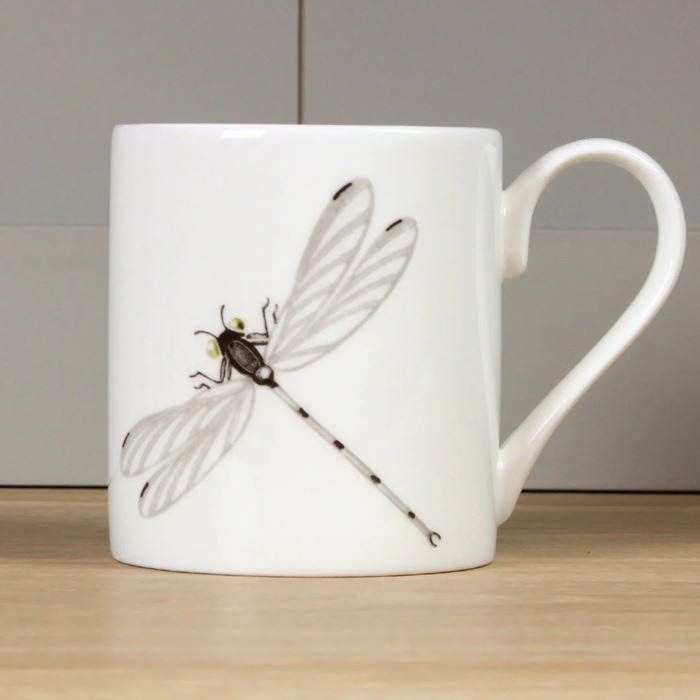 Dragonfly mug by holly lasseter shows a water colour drawing of a dragonfly in grey and black on a white mug