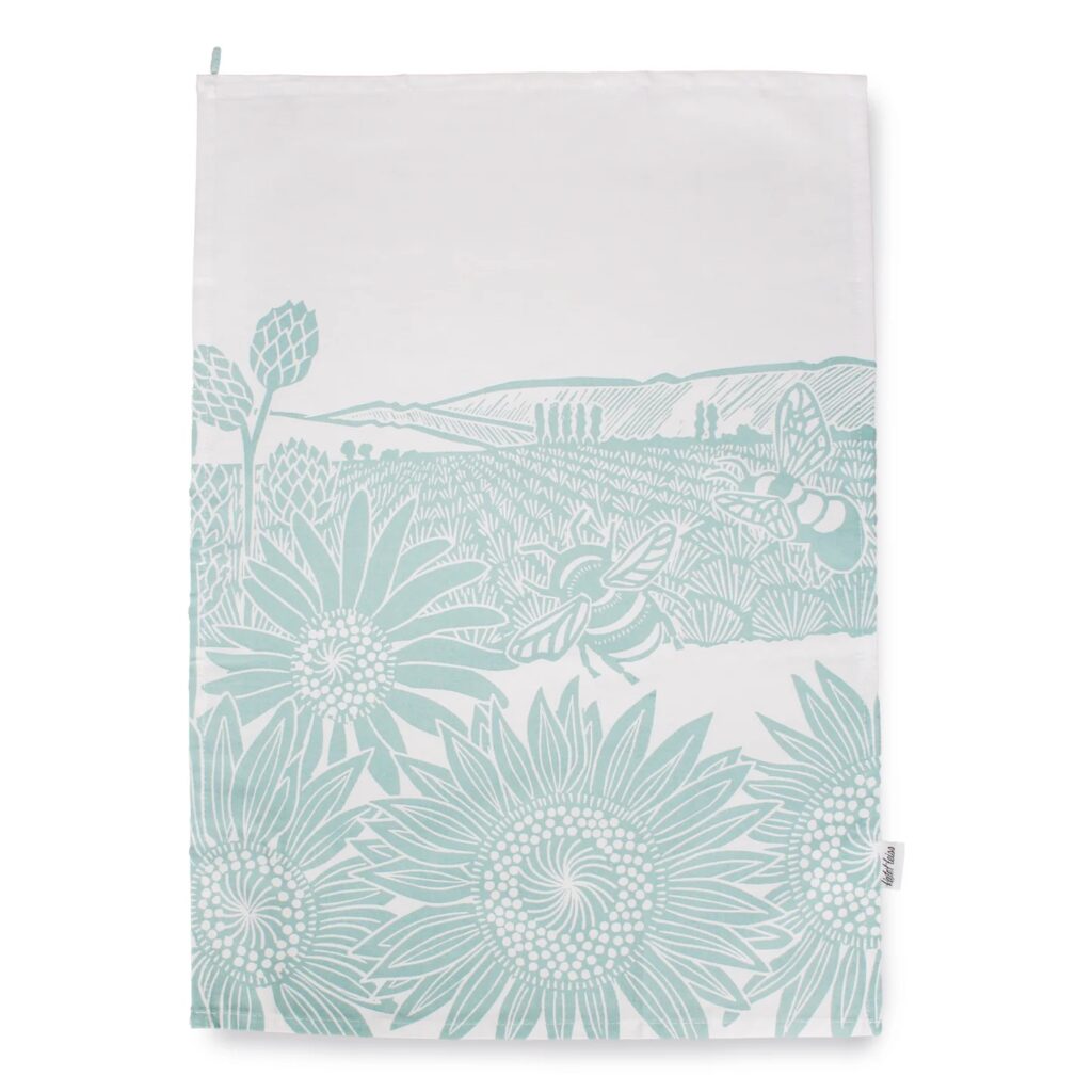 Kate Heiss Sunflowers and Bees Tea Towel in Powder Blue shows a white and blue tea towel with an illustration of bees and sunflowers against fields