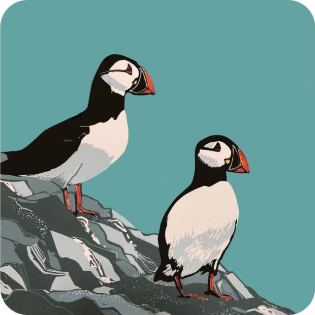 Puffins Placemat by Sue Welfare shows two black and white birds with colourful bills standing on the edge of a cliff against a blue background