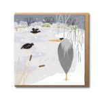 Heron and Frozen Pond Greetings Card by Rachel Hudson shows a grey heron standing on the side of a frozen pond whilst two small black birds peck on the ice